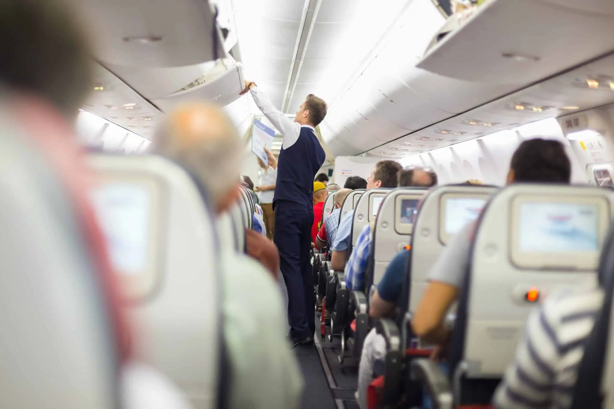the interior of an airplane with passengers in seats and a flight attendant closing overhead luggage compartments
